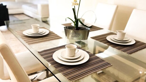 A modern dining area with a glass table, white chairs, and a metal bucket labeled "FLOWERS & GARDEN" containing orchids. - 5BR Pool+Hot Tub Home Kissimmee Near Disney WDW (Kissimmee)