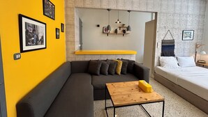 Ein modernes Wohnzimmer mit Sofa, einem kleinen Holztisch, einem Bett und einer Küchenzeile mit einer Pendelleuchte.