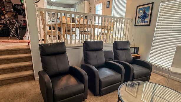 A living room with a glass coffee table, three black leather chairs, a staircase with a white railing, and a kitchen in the background. - Nice 2 bedroom home with backyard only 15 mins outside of Atlanta. (Detroit)