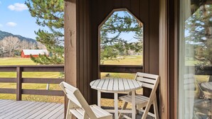 Una terraza de madera con una mesa y dos sillas, con vista a un área verde con árboles y un granero rojo.