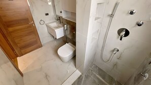 Une salle de bain moderne avec un sol en marbre, une porte en bois, un lavabo blanc, des toilettes et une douche dotée de plusieurs commandes.