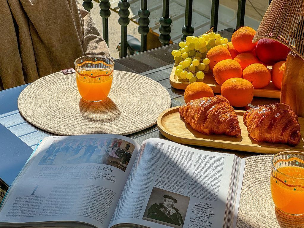 Avoimena oleva kirja, lasi appelsiinimehua, lautanen, jossa on appelsiineja ja croissantteja, ja rottinkimatto pöydällä.