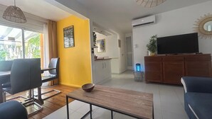A modern living room with a sofa, a coffee table, and a TV on a wooden cabinet. There is a dining area with a table and chairs, and a kitchen in the background. - Cocon Tropical (Sainte-Anne)