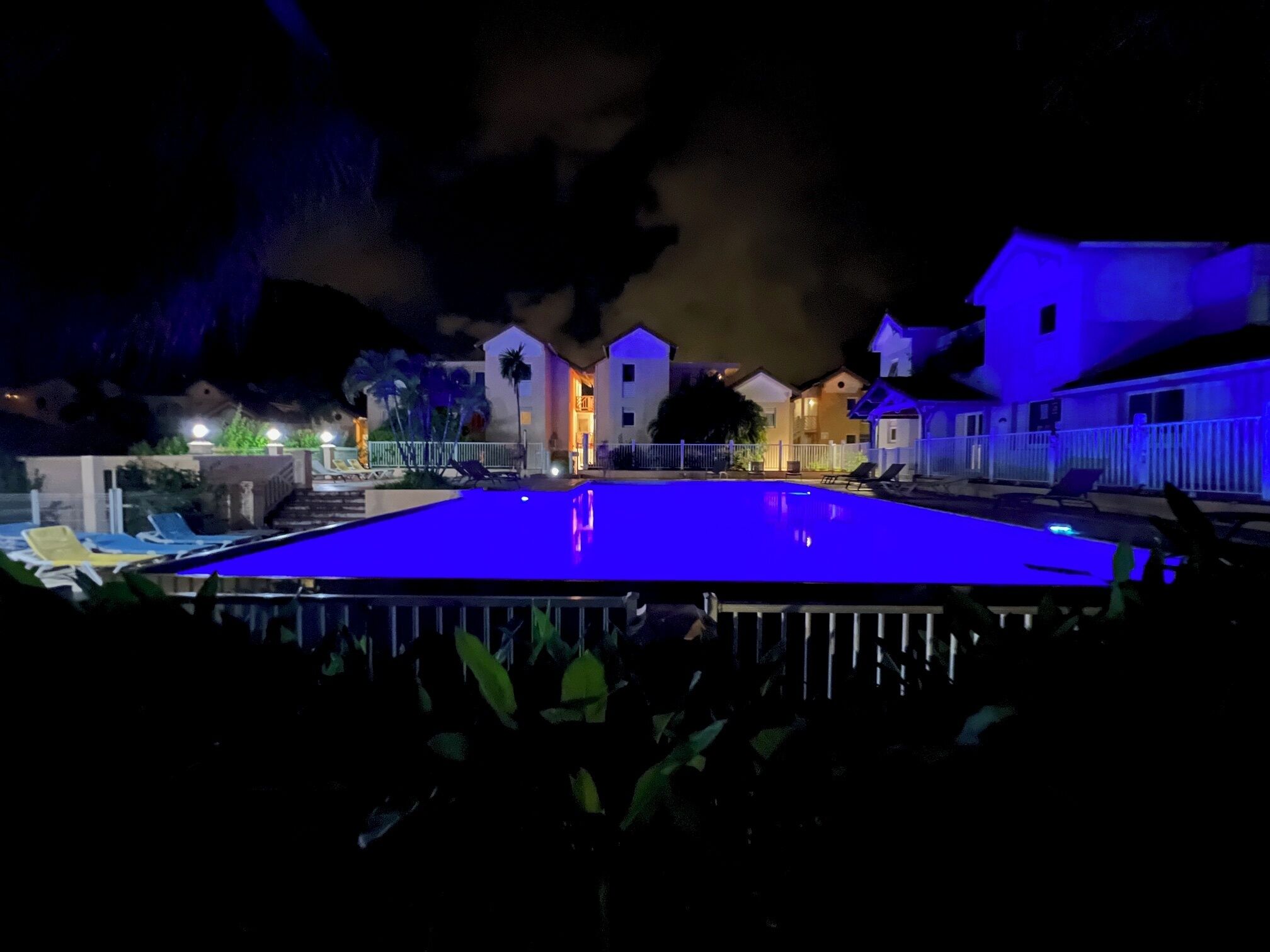 A swimming pool with a blue tint, surrounded by a white fence, and adjacent buildings with illuminated windows.