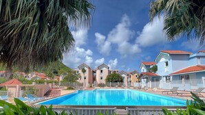 A swimming pool surrounded by lounge chairs and palm trees, with buildings in the background. - Cocon Tropical (Sainte-Anne)