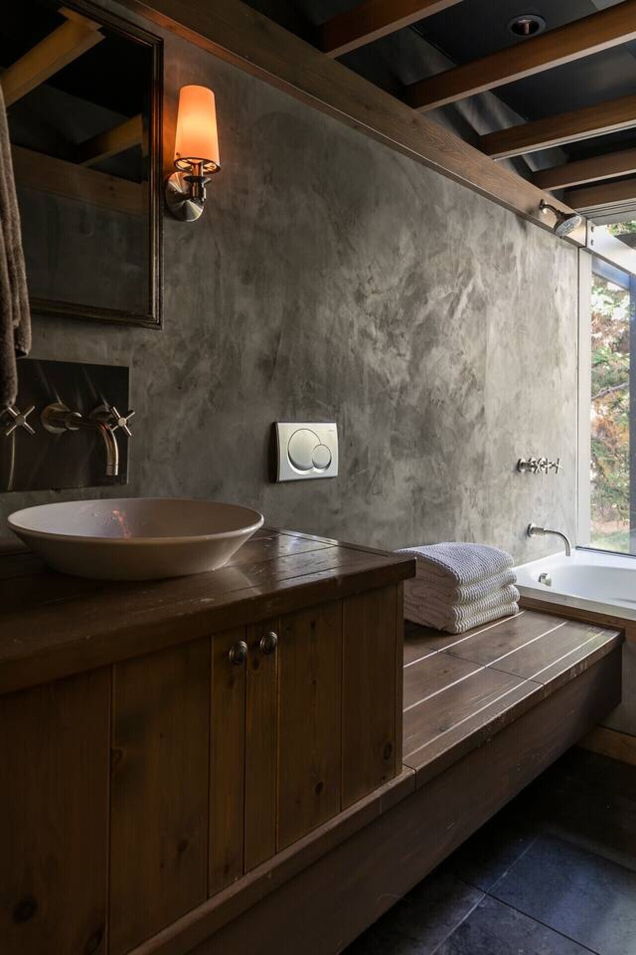 A modern bathroom with a wooden vanity, a round sink, a built-in shower, and a large mirror with a wall-mounted light.