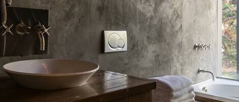 A modern bathroom with a wooden vanity, a round sink, a built-in shower, and a large mirror with a wall-mounted light.