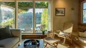 Una stanza accogliente con un divano, due poltrone in vimini, un tavolino con un vassoio e vista su un lago e una foresta attraverso ampie finestre.