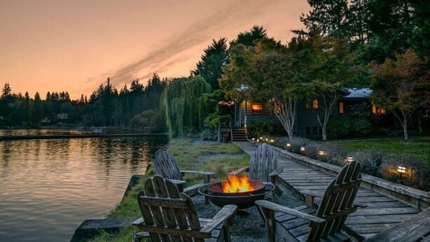 Una terrazza sul lago con un braciere e quattro poltrone Adirondack, circondata da un percorso in legno con una casa sullo sfondo.