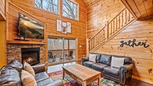 A cozy cabin interior with a stone fireplace, leather sofas, a wooden coffee table, and a staircase. - Heated Pool (seasonal), Hot Tub, Game Room, & Mountain Views – Sleeps 12 (Sevierville)
