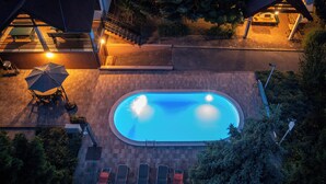 Eine Vogelperspektive auf ein Schwimmbecken mit Liegestühlen und einem Tisch, umgeben von Bäumen und einem Gebäude mit einer Treppe.