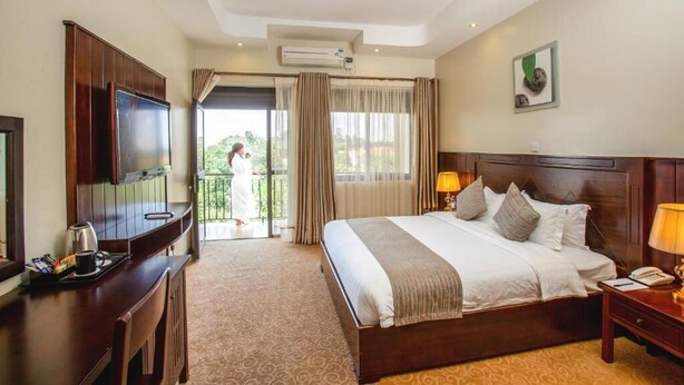 A hotel room with a large bed, a TV, a desk with a coffee maker, and a balcony with a view of greenery. - 6Cape Guest House Kawanda  (Kampala)