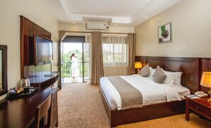 A hotel room with a large bed, a TV, a desk with a coffee maker, and a balcony with a view of greenery. - 6Cape Guest House Kawanda  (Kampala)