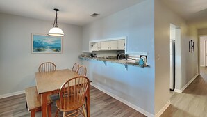 A dining area with a wooden table and chairs, a hanging light fixture, and a kitchen with white cabinets and a countertop. - Ocean Echo #303 (Gulf Shores)