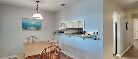 Un comedor con mesa y sillas de madera, un lámpara colgante, y una cocina con armarios blancos y encimera.