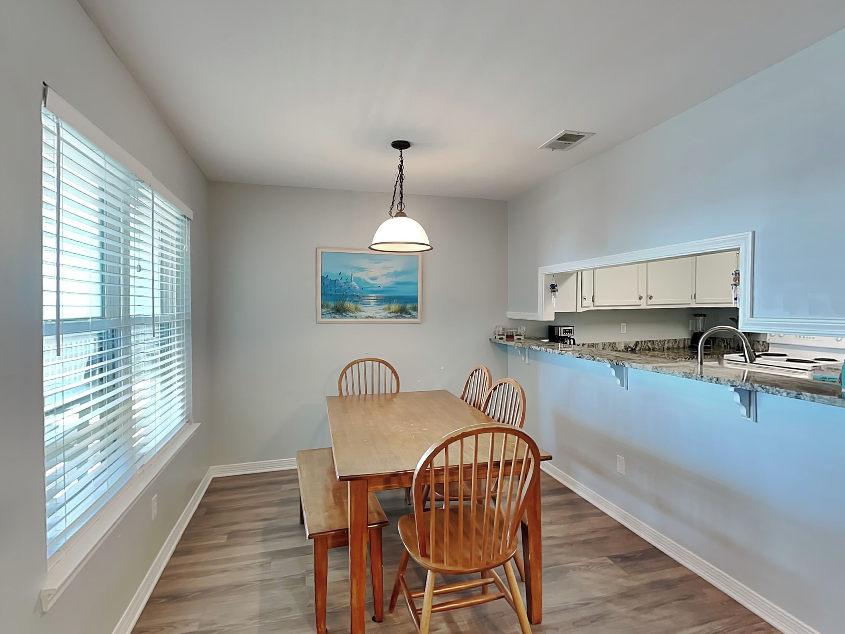 A dining area with a wooden table and chairs, a pendant light, a painting on the wall, and a kitchen with white cabinets and a marble countertop.