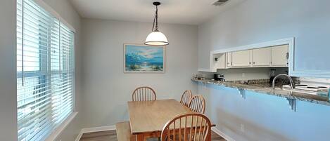 A dining area with a wooden table and chairs, a pendant light, a painting on the wall, and a kitchen with white cabinets and a marble countertop.