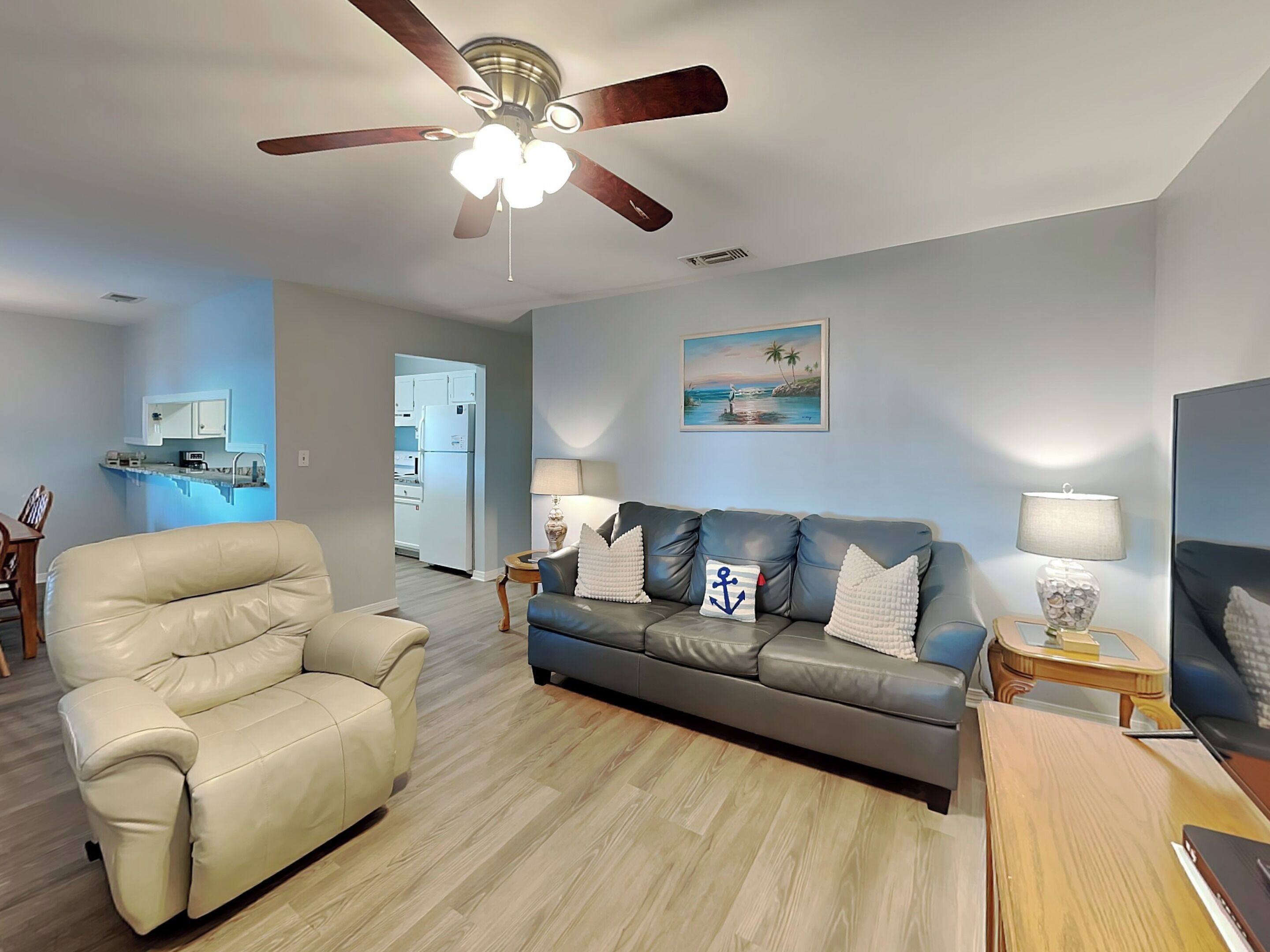 Una sala de estar con un sillón de cuero color beige, un sofá gris con almohadones de temática náutica, una mesa de centro de madera y un ventilador de techo.