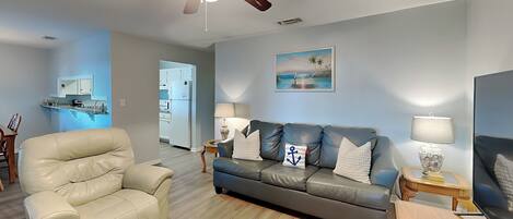 Una sala de estar con un sillón de cuero color beige, un sofá gris con almohadones de temática náutica, una mesa de centro de madera y un ventilador de techo.