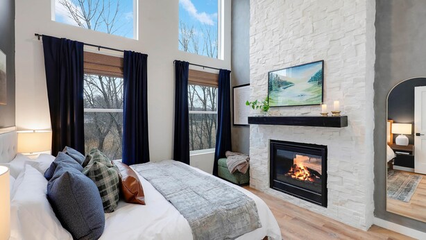 Un dormitorio con una cama grande, una chimenea y vistas a árboles desde las ventanas.