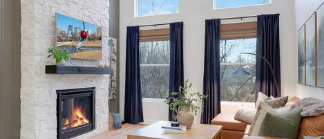 A spacious living room with a fireplace, a ceiling fan, and large windows offering a view of the outdoors.