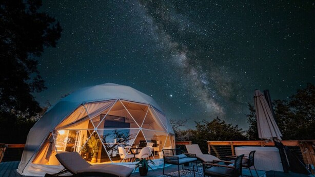 Una tienda en forma de domo geodésico con un área de patio que incluye sillones y una mesa de comedor, frente a un cielo nocturno con estrellas visibles y la Vía Láctea.