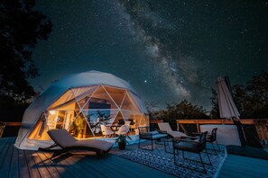 Una tienda en forma de domo geodésico con un área de patio que incluye sillones y una mesa de comedor, frente a un cielo nocturno con estrellas visibles y la Vía Láctea.