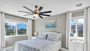 Une chambre à coucher comprenant un lit, un ventilateur de plafond, une table de chevet, une fenêtre avec des rideaux et une vue sur l’extérieur.