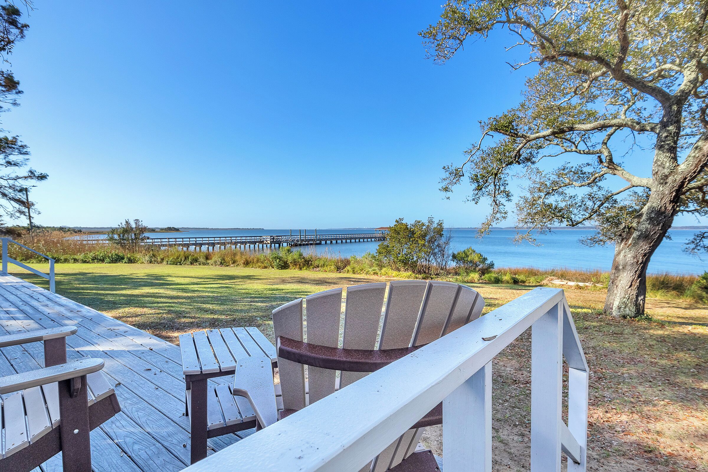 A wooden deck with picnic tables and a Adirondack chair overlooking a body of water with a pier in the distance.