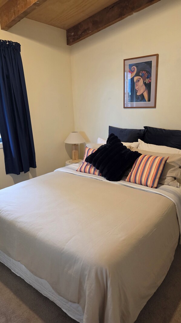 Cama bem arrumada com lençóis brancos, manta preta e almofadas listradas. Há uma obra de arte emoldurada na parede acima da cama.