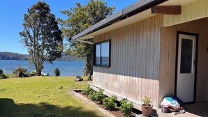 A wooden cabin with a large window overlooking a lake, surrounded by a grassy yard and a few potted plants. - Lake Rotoiti - Absolute Lakefront (Rotoiti Forest)
