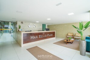 Una zona de recepción con un mostrador de madera con la etiqueta "MAR BELLO PLAZA", un área de descanso con sillas y una mesa de centro, y una planta grande en maceta.