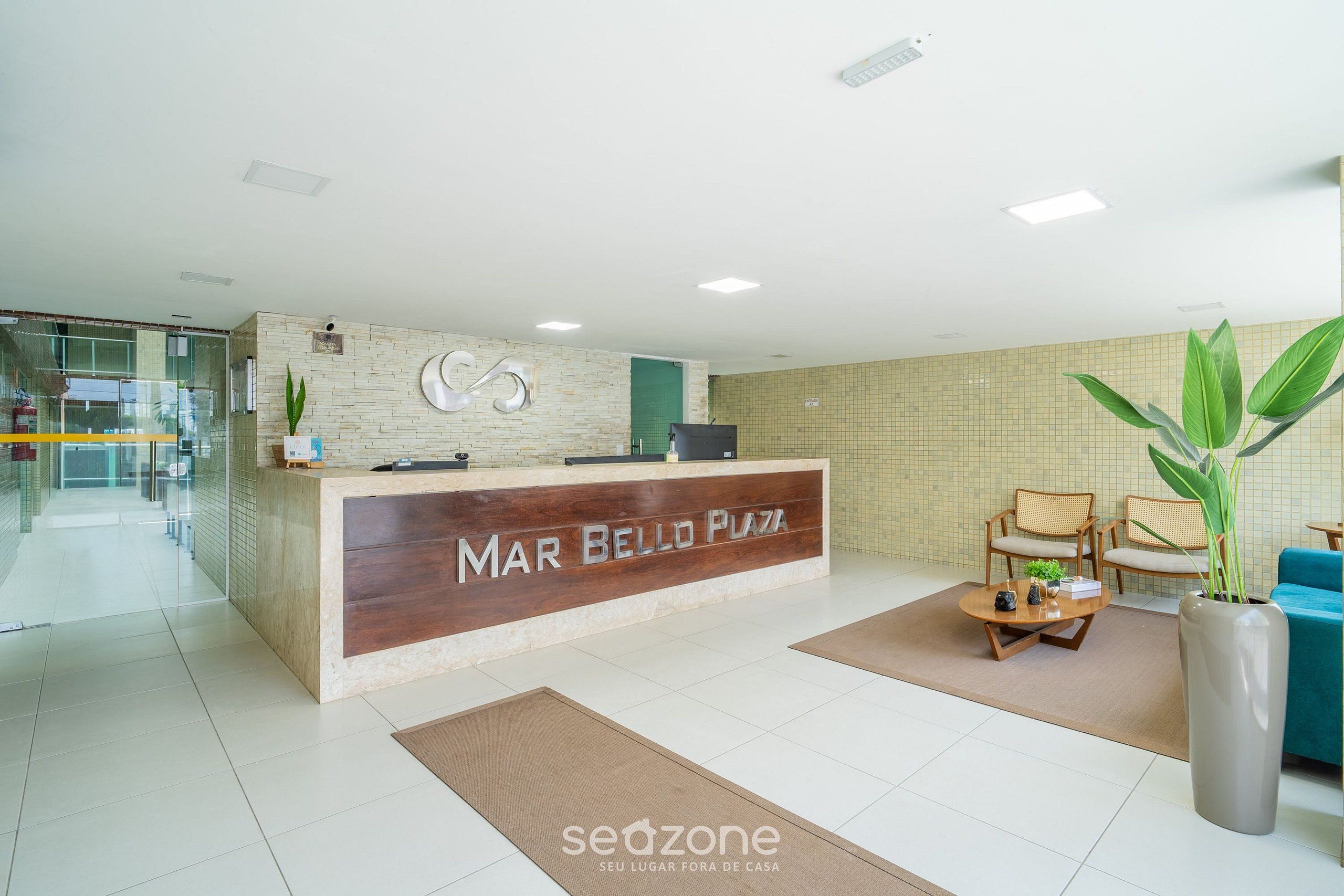 A reception area with a wooden reception desk labeled "MAR BELLO PLAZA," a seating area with chairs and a coffee table, and a large potted plant.