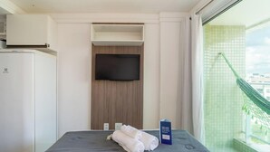 A hotel room with a bed, a flat-screen TV mounted on a wooden panel, a minibar, and a view of a green outdoor area. - Your Complete Stay With a Rooftop Pool, 40m From the Beach in Cabedelo Cmzs (Cabedelo)