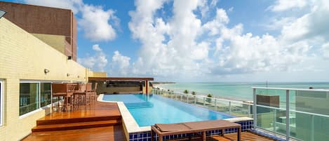 A rooftop pool with a view of the ocean, wooden deck, and outdoor seating.