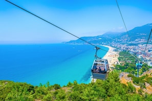 A cable car moving over a coastal area with a beach and cityscape in the background. - Apartment 2+1 Qoople  Elize 29 Boho Style in Premium location (Alanya)