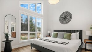A modern bedroom with a large bed, a wall-mounted circular artwork, and a large window offering a view of the outdoors. - Chalet Grande Ourse | Slope-Side | Private Hot Tub (Panorama)