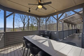 A covered outdoor deck with a long table and chairs, a ceiling fan, and a view of the surrounding trees and hills. - Paxwood, Luxury Home for 16 (Dawsonville)