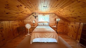 Een knus slaapkamer op zolder, met een houten schuine plafond, een bed met een witte deken, een ventilator en een nachtkastje met een lamp.