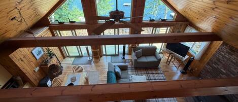 A high-ceilinged wooden cabin with a loft bedroom, a living area with a sofa and TV, and large windows offering views of the outdoors.