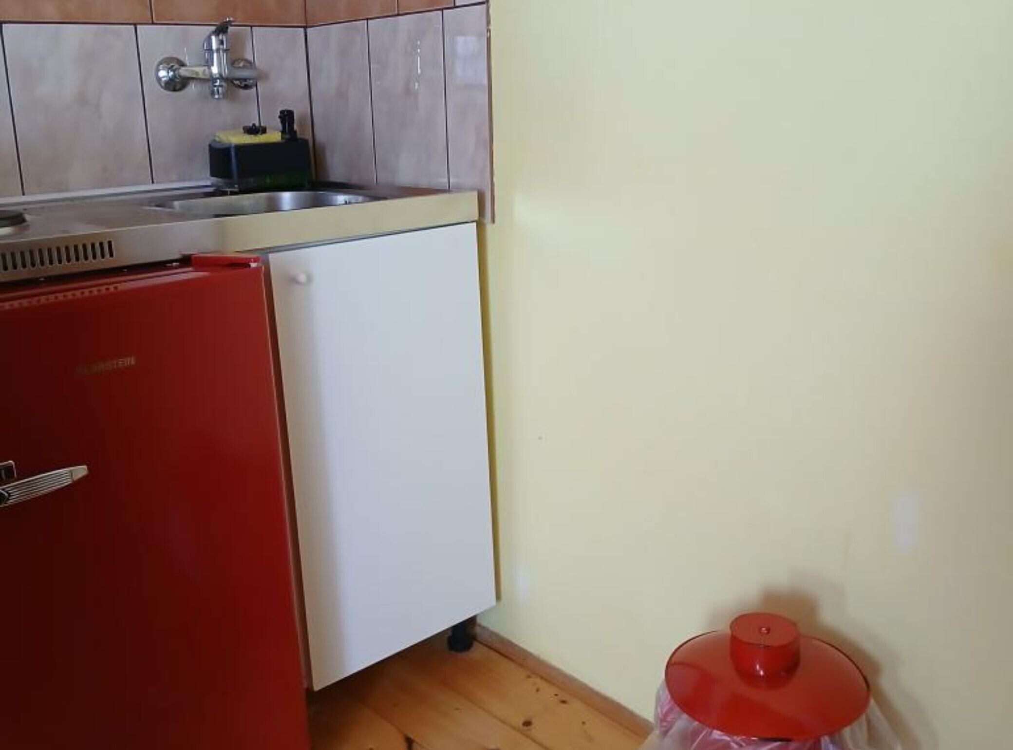 A compact kitchen with a sink, a small refrigerator, and a red trash bin.