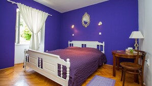 Un dormitorio con cama, ventana con cortinas blancas, mesita de noche con lámpara y espejo en la pared.