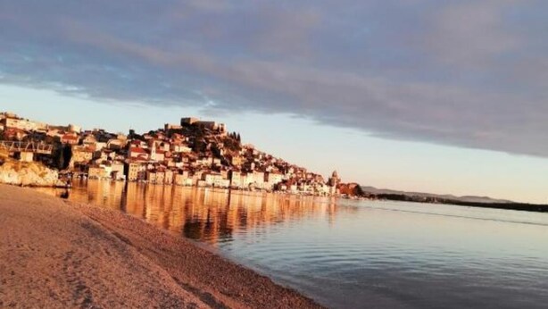 Un pueblo costero con edificios a lo largo del litoral, una playa arenosa y agua tranquila que refleja las construcciones.