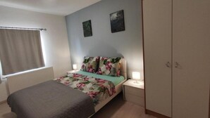 Un dormitorio con cama, dos mesitas de noche, un armario, una ventana con cortinas y dos obras de arte colgadas en la pared.