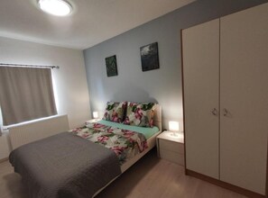 Ein Schlafzimmer mit einem Bett, zwei Nachttischen, einem Kleiderschrank, einem Fenster mit Vorhängen und zwei Wandbildern.