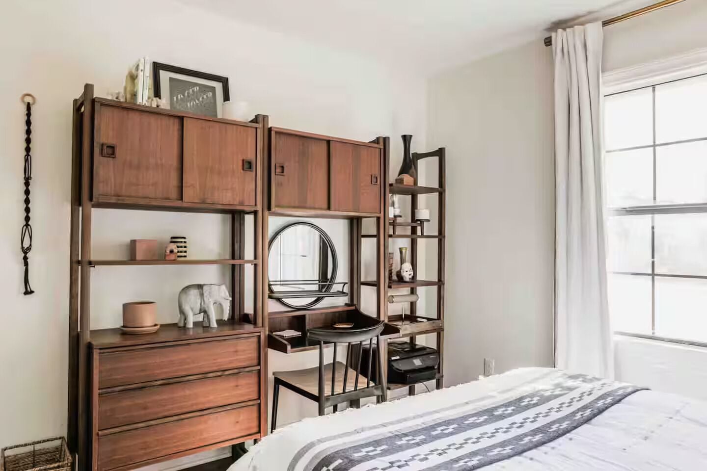 A bedroom with a bed, wooden wardrobe, shelves, a mirror, and a window with curtains.