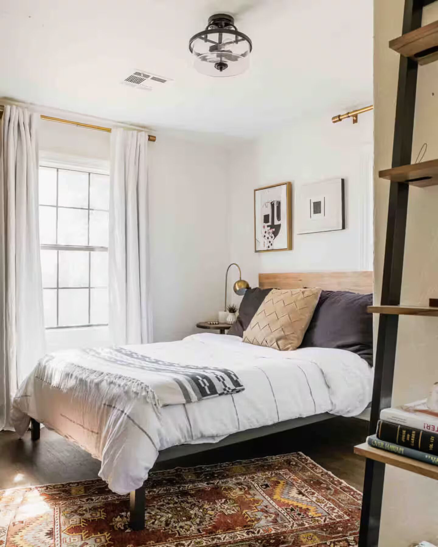 Um quarto com cama, mesa de cabeceira, luminária, janela com cortinas e ventilador de teto.