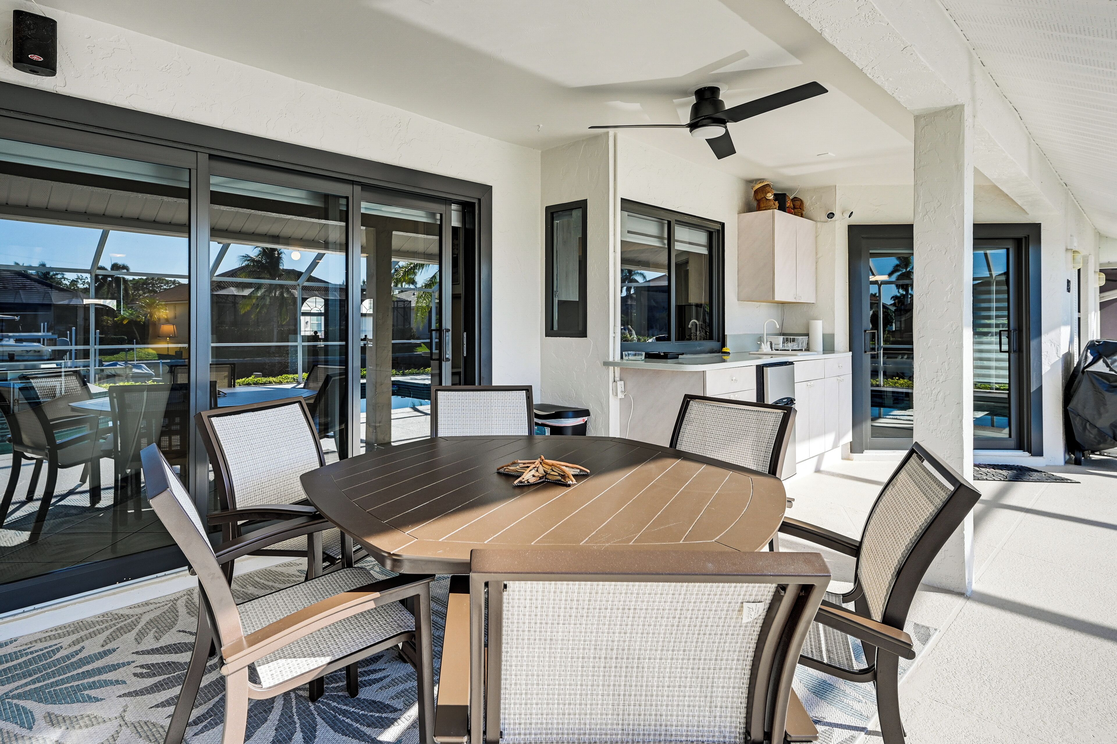 A modern outdoor dining area with a large table and chairs, a ceiling fan, and a view of the surrounding landscape.