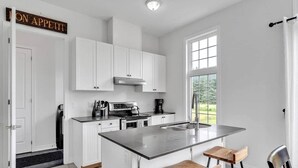 A modern kitchen with white cabinetry, a central island with bar stools, a stove, and a large window providing natural light. - Spacious & New Prince Edward County Family Home (Quinte West)