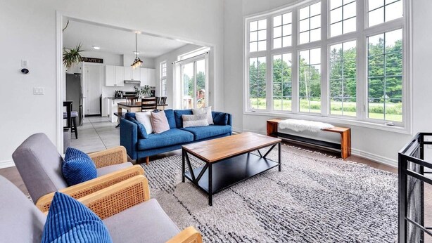 A spacious living room with a blue sofa, a wooden coffee table, and large windows offering a view of greenery. - Spacious & New Prince Edward County Family Home (Quinte West)